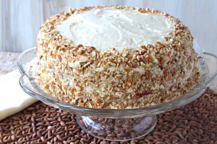 An entire banana poppy seed cake covered in pecans on a glass cake plate.