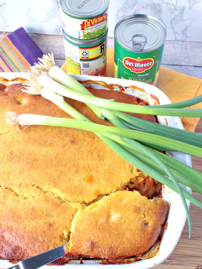 A vertical overhead picture of a chili mac casserole with cornbread crust in a casserole dish with whole scallions and a serving spoon.