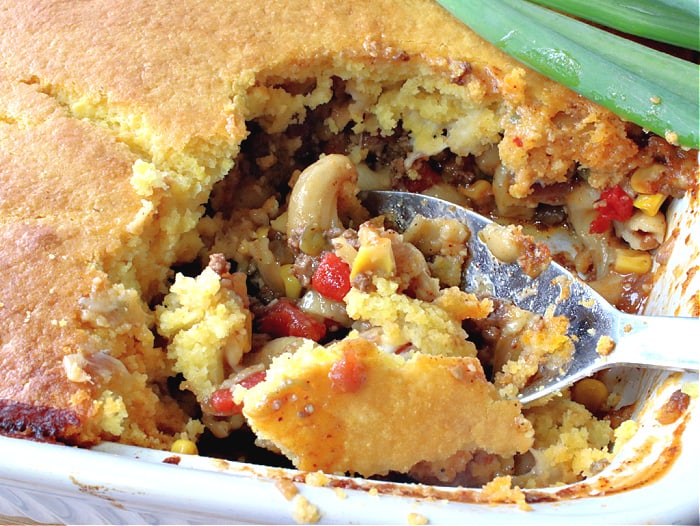 Closeup overhead photo of a chili mac with a cornbread crust casserole with corn, red peppers, macaroni and a large serving spoon.