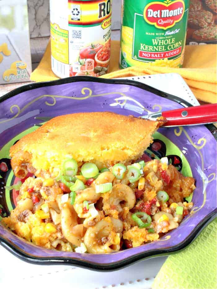 A vertical closeup image of chili mac casserole in a purple bowl with scallions, corn, and ground beef and can corn and tomatoes in the background.
