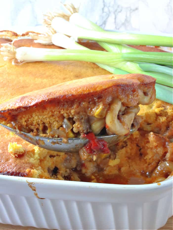 A vertical photo of a serving spoon with a bit helping of chili mac with cornbread crust out of a casserole dish.