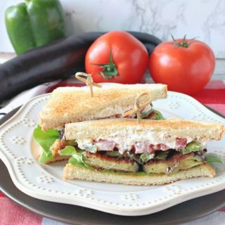 A fried eggplant sandwich on a white plate on a red and white tablecloth with a bell pepper, eggplant, and tomatoes in the background.
