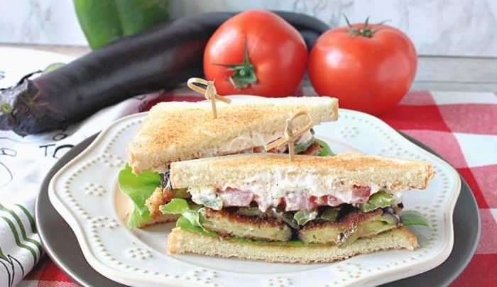 A fried eggplant sandwich on a white plate on a red and white tablecloth with a bell pepper, eggplant, and tomatoes in the background.