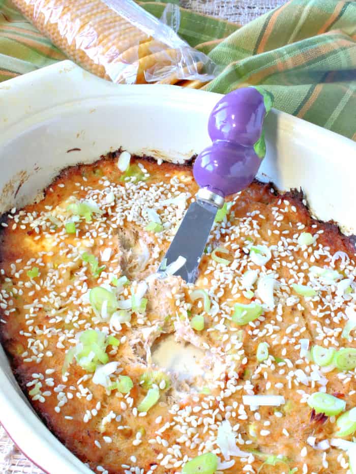 A baked crab rangoon spread with a cute eggplant appetizer knife in the center and scallions and sesame seeds sprinkled on top.