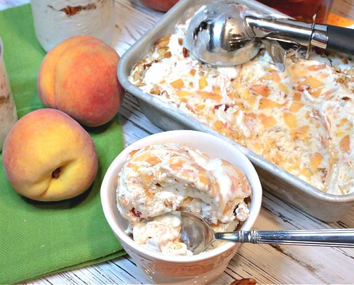 An overhead photo of a dish of no-churn peach ice cream and a spoon along with a loaf pan, a green napkin, and fresh peaches in the background.