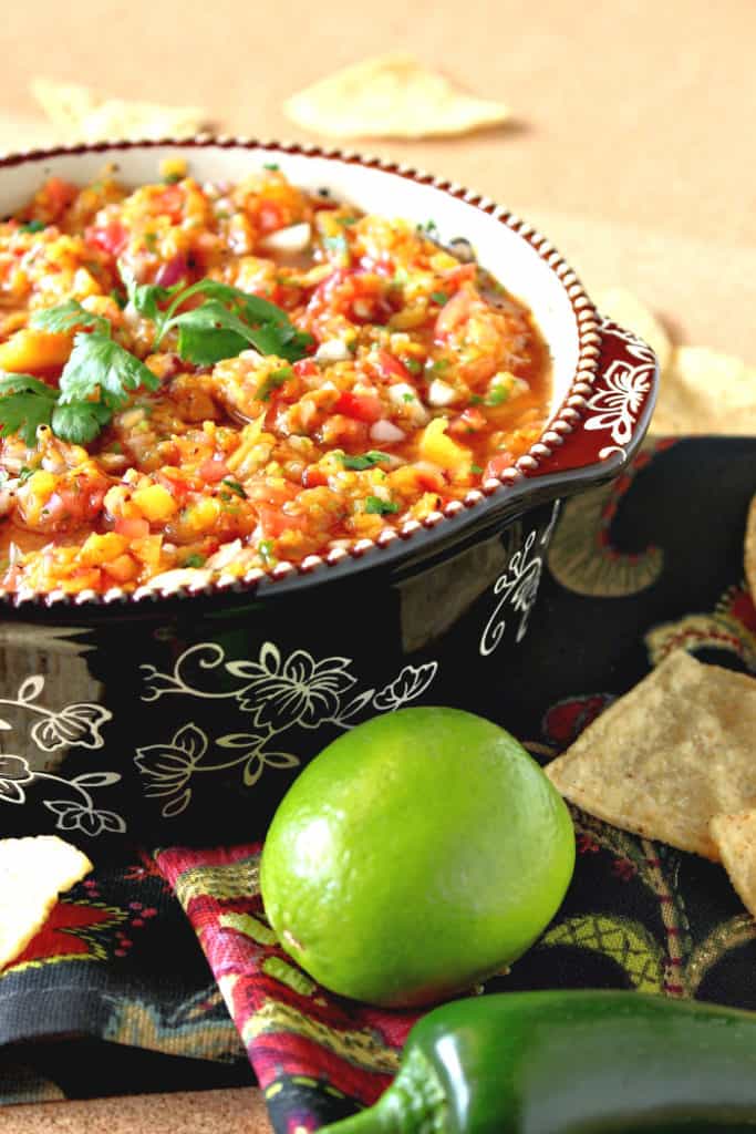 A vertical closeup photo of grilled peach salsa in a bowl with fresh cilantro and a lime and jalapeno pepper in the foreground.