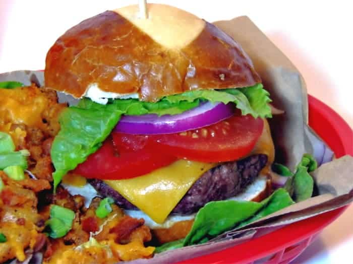 Closeup photo of a ground short rib burger with Italian sausage, a pretzel bun, cheese, tomato, and lettuce.