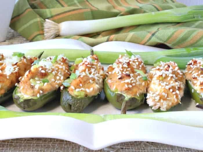 A closeup horizontal photo of jalapeno poppers stuffed with crab rangoon on a white rectangle platter topped with chopped scallions and a green and gold napkin in the background. How to make jalapeno poppers with crab rangoon filling