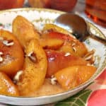An offset photo of a bowl filled with peaches foster with chopped pecans over top.