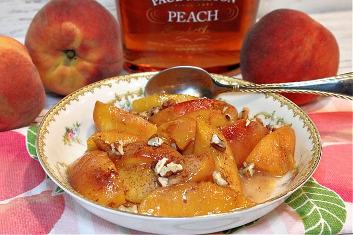 A pretty china bowl filled with peaches foster and topped with chopped pecans. Fresh peaches and peach brandy are in the background