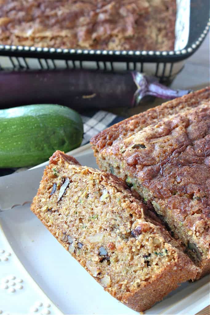 A vertical closeup of a sliced eggplant zucchini bread on a plate with a a whole loaf in a baking pan in the background.