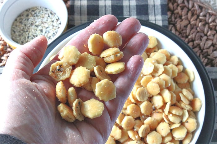 A closeup of a hand holding a bunch of everything seasoned oyster crackers in the foreground and a bowl with the crackers in the background.