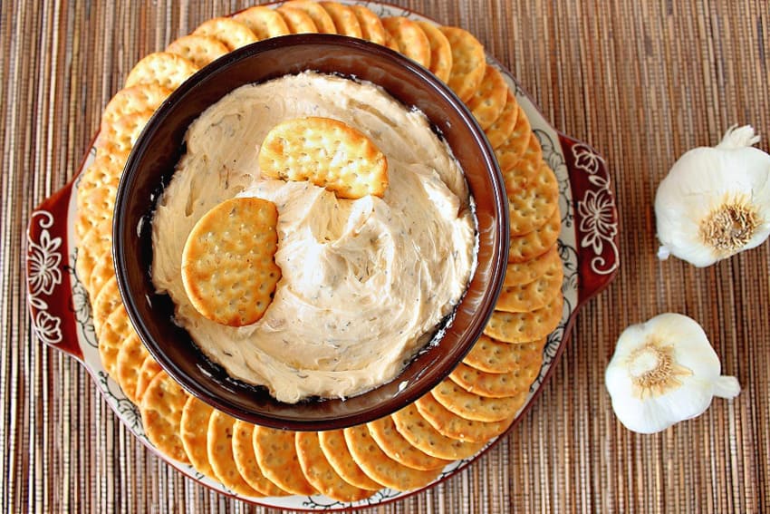 An overhead photo of a brown bowl filled with roasted garlic dip on a platter surrounded by crackers and garlic bulbs on the side.