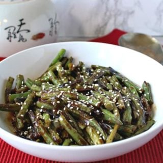 A white bowl of blistered green beans with miso and sesame on a red table cloth with a serving spoon.
