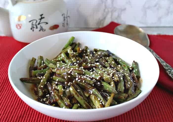 A white bowl of blistered green beans with miso and sesame on a red table cloth with a serving spoon.