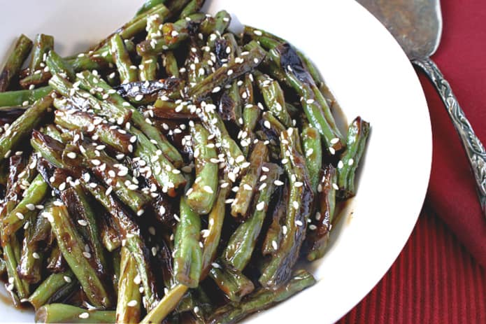 A closeup photo of blistered green beans with miso sauce and sesame seeds on a red tablecloth with a serving spoon on the side.