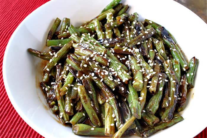 An offset photo of a bowl of blistered green beans with sesame seeds and miso sauce on a red background.