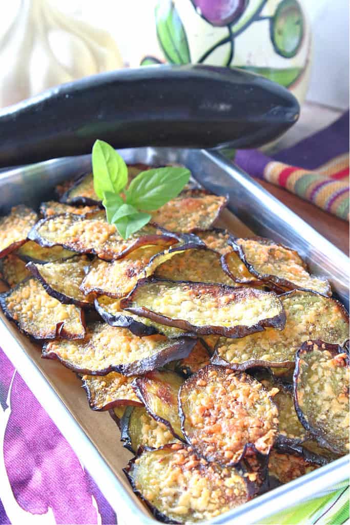 A vertical closeup picture of a small baking dish filled with crispy baked eggplant chips with fresh basil as a garnish, and an olive oil bottle in the background.