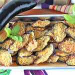 A small baking tray filled with crispy baked eggplant chips with an whole eggplant and fresh basil as garnish