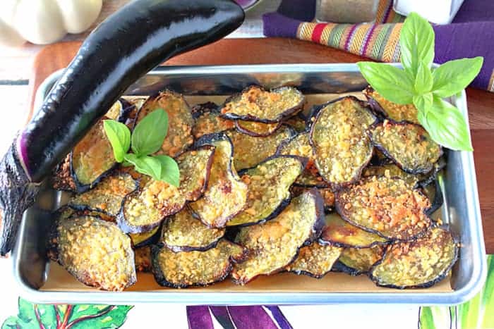 A small baking tray filled with crispy baked eggplant chips with an whole eggplant and fresh basil as garnish