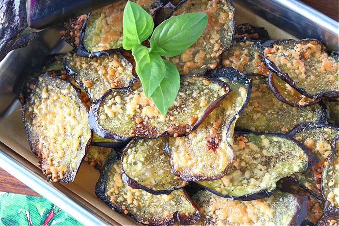 An overhead closeup photo of a pile of crispy baked eggplant chips with Parmesan cheese and a fresh basil sprig.