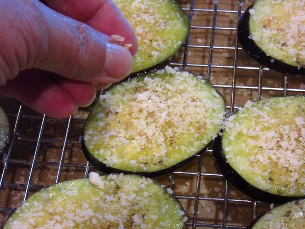A hand sprinkling Parmesan cheese onto eggplant slices.