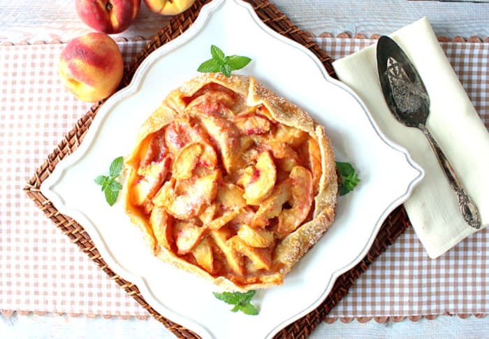 An overhead picture of a peach crostata on a square plate on a tan and white checked tablecloth with mint and fresh peaches for garnish