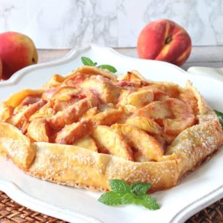 A fresh peach crostata on a square white platter with mint garnish and fresh peaches in the background.