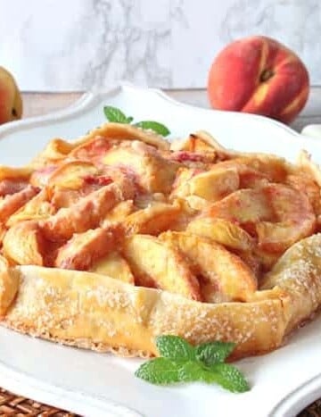 A fresh peach crostata on a square white platter with mint garnish and fresh peaches in the background.