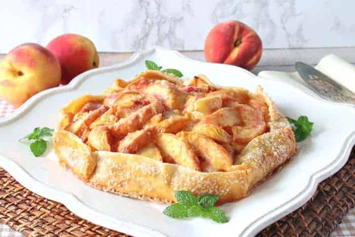 A fresh peach crostata on a square white platter with mint garnish and fresh peaches in the background.