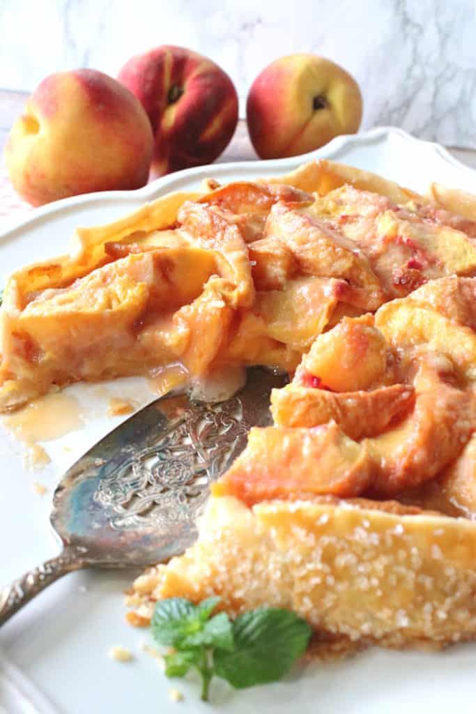 A vertical closeup photo of a peach crostata on a white platter with a slice taken out and a pie server and fresh mint.