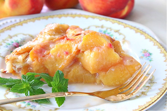 A very closeup photo of a slice of fresh peach crostata on a pretty china plate with a fork, and some mint sprigs for garnish.