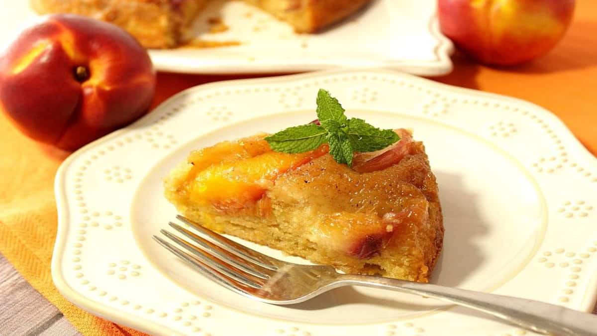 A slice of Nectarine Upside Down Cake on a white plate with a fork and a nectarine in the background.