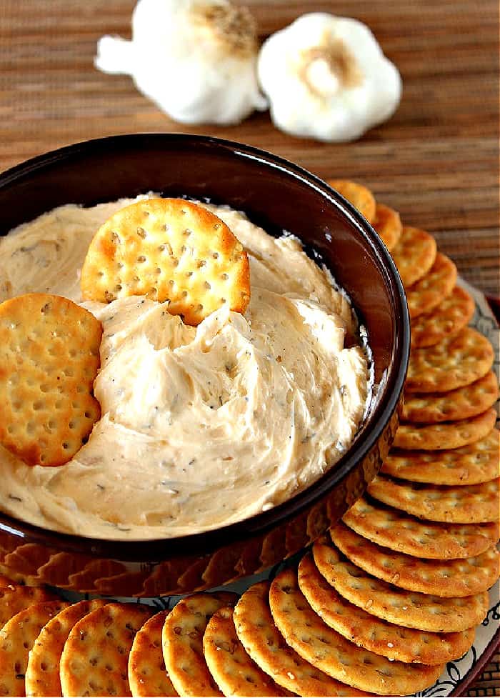 A vertical closeup photo of a bowl filled with roasted garlic dip with crackers around the outside of the bowl and inside the dip.