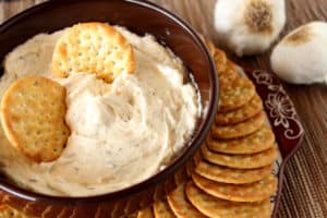 A brown bowl filled with roasted garlic dip with two crackers in the dip.