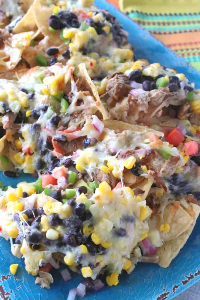 A vertical closeup of Loaded Pulled Pork Nachos on a blue platter with black beans, onions, and corn.
