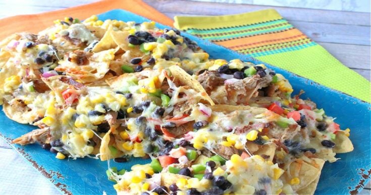 A colorful photo with loaded pulled pork nachos on a blue platter with a lime green and orange napkin in the background