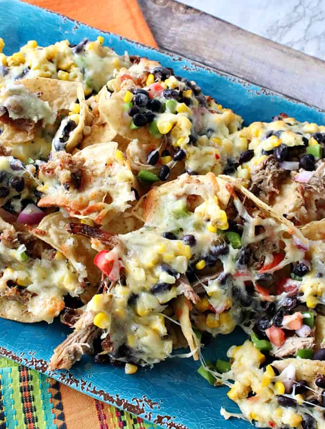 A vertical closeup of Loaded Pulled Pork Nachos on an aqua blue plate with corn, green peppers, tomato, and black beans