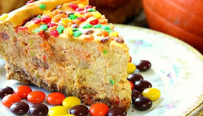 A super closeup photo of a slice of Reese's Pieces Pumpkin Cheesecake on a plate with Reese's pieces candy and fall sprinkles.