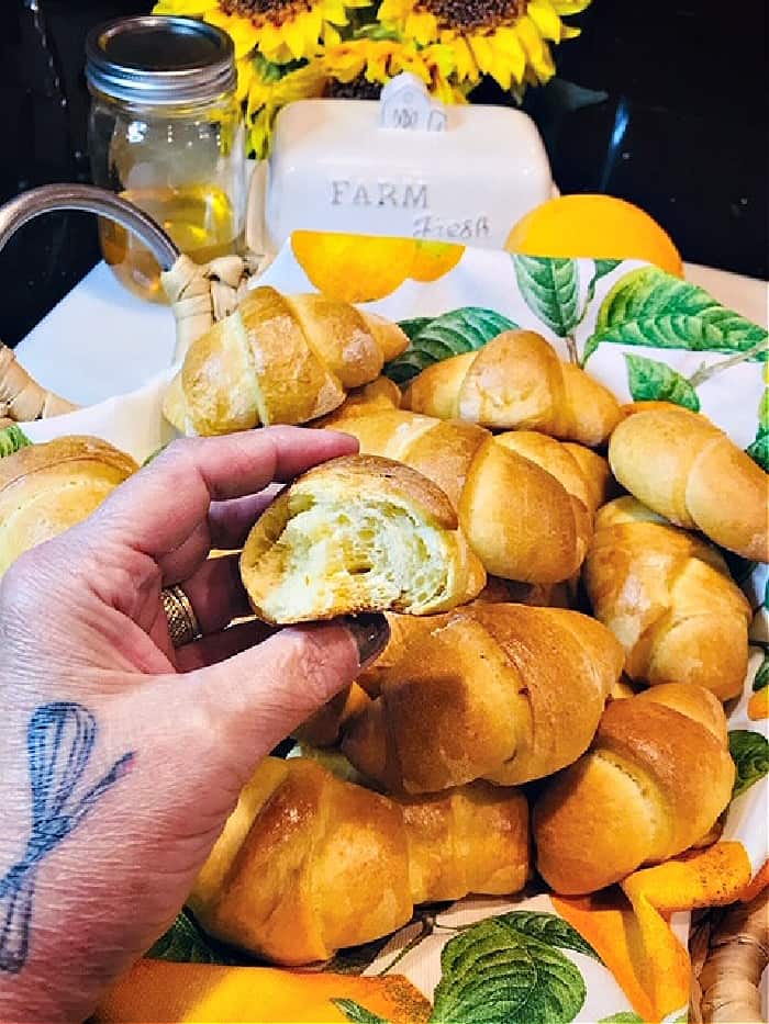 A closeup vertical photo of a hand holding a honey orange crescent roll with a full basket in the background