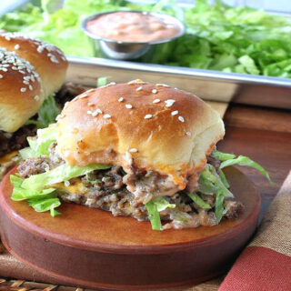 A big mac slider on a wooden pedestal with sliders in the background and a tray of lettuce and secret sauce