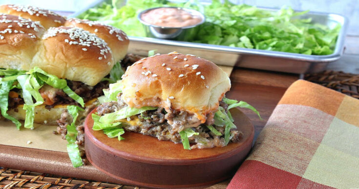 A big mac slider on a wooden pedestal with sliders in the background and a tray of lettuce and secret sauce