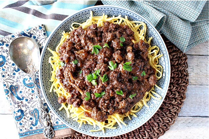 Overhead photo of a blue bowl filled with ground beef sauerbraten goulash with a serving spoon and blue napkins.