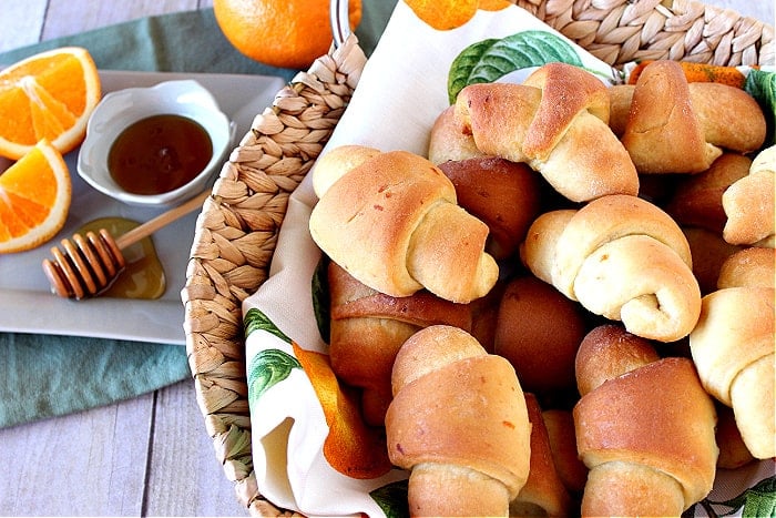 A horizontal photo of a basket filled with honey orange crescent rolls on the right and a dish on honey and some fresh oranges on the left.