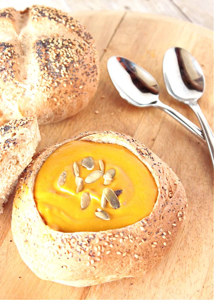 A vertical photo of a seeded bread bowl filled with Roasted Butternut Squash Soup along with two spoons in the background and another seeded loaf of bread.