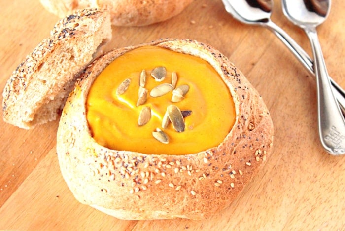 A seeded bread bowl filled with Roasted Butternut Squash Soup, two spoons, and pepita seeds as garnish