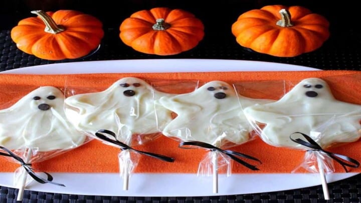 A lineup of white chocolate ghost pops on an orange napkin with mini pumpkins in the background