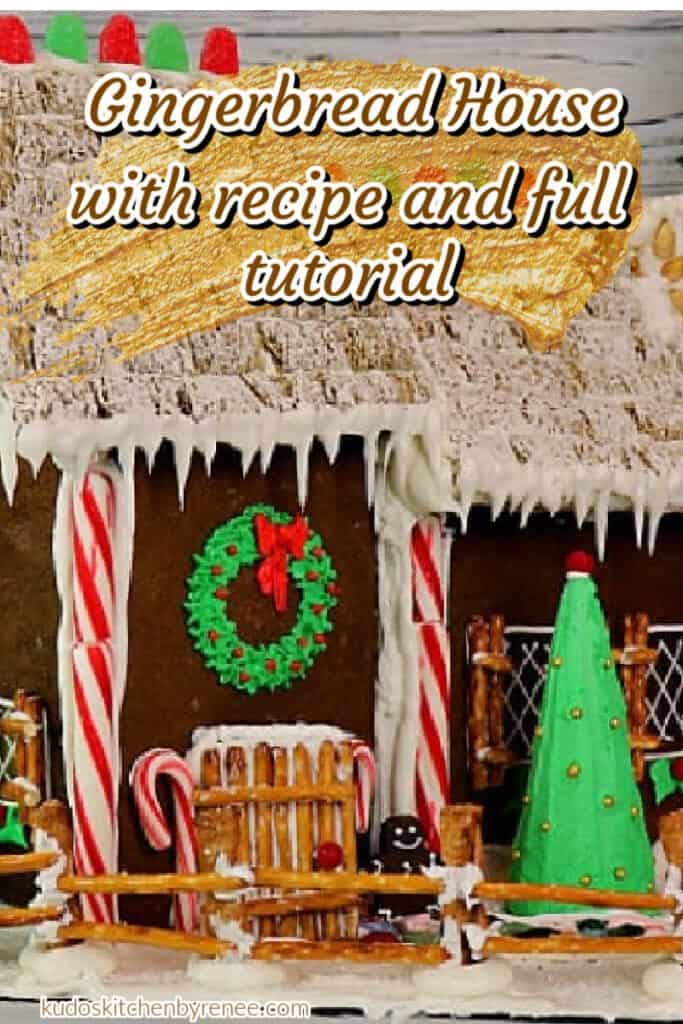 A closeup vertical image of a Gingerbread House Recipe with pretzel sticks, peppermint sticks, royal icing, and a green wreath above the door with a title text overlay graphic.