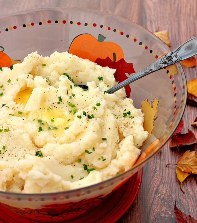 A vertical closeup of mashed celery root potatoes in a colorful glass bowl with melted butter, pepper, and minced chives on top.