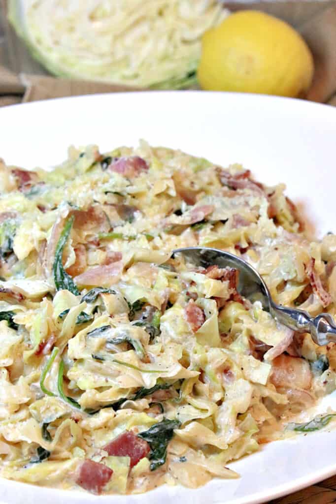 A closeup vertical image of a bowl of Creamed Cabbage and Spinach with Bacon along with a wedge of cabbage and a lemon in the background